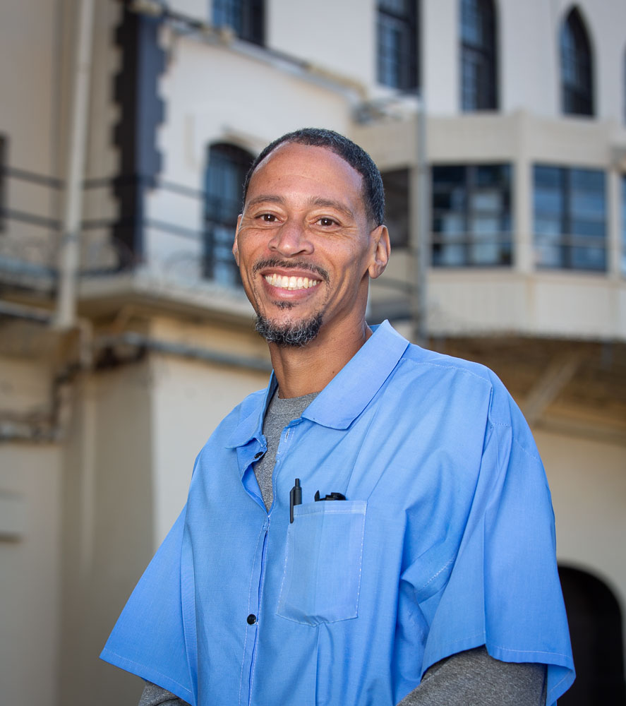Black man with light blue shirt over a gray long sleeve.