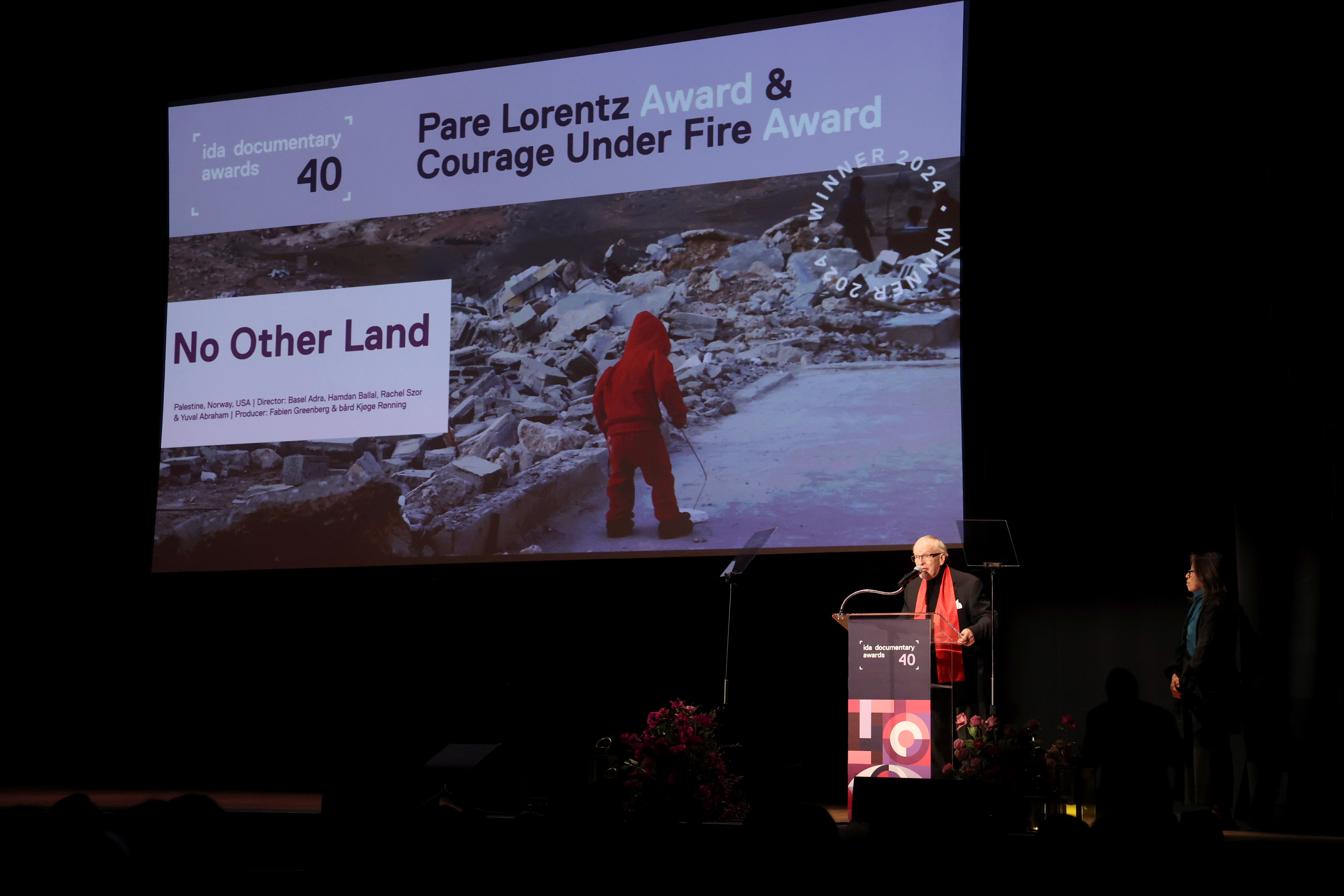 A person at a podium speaks to a crowd with a large screen with a still from "No Other Land"
