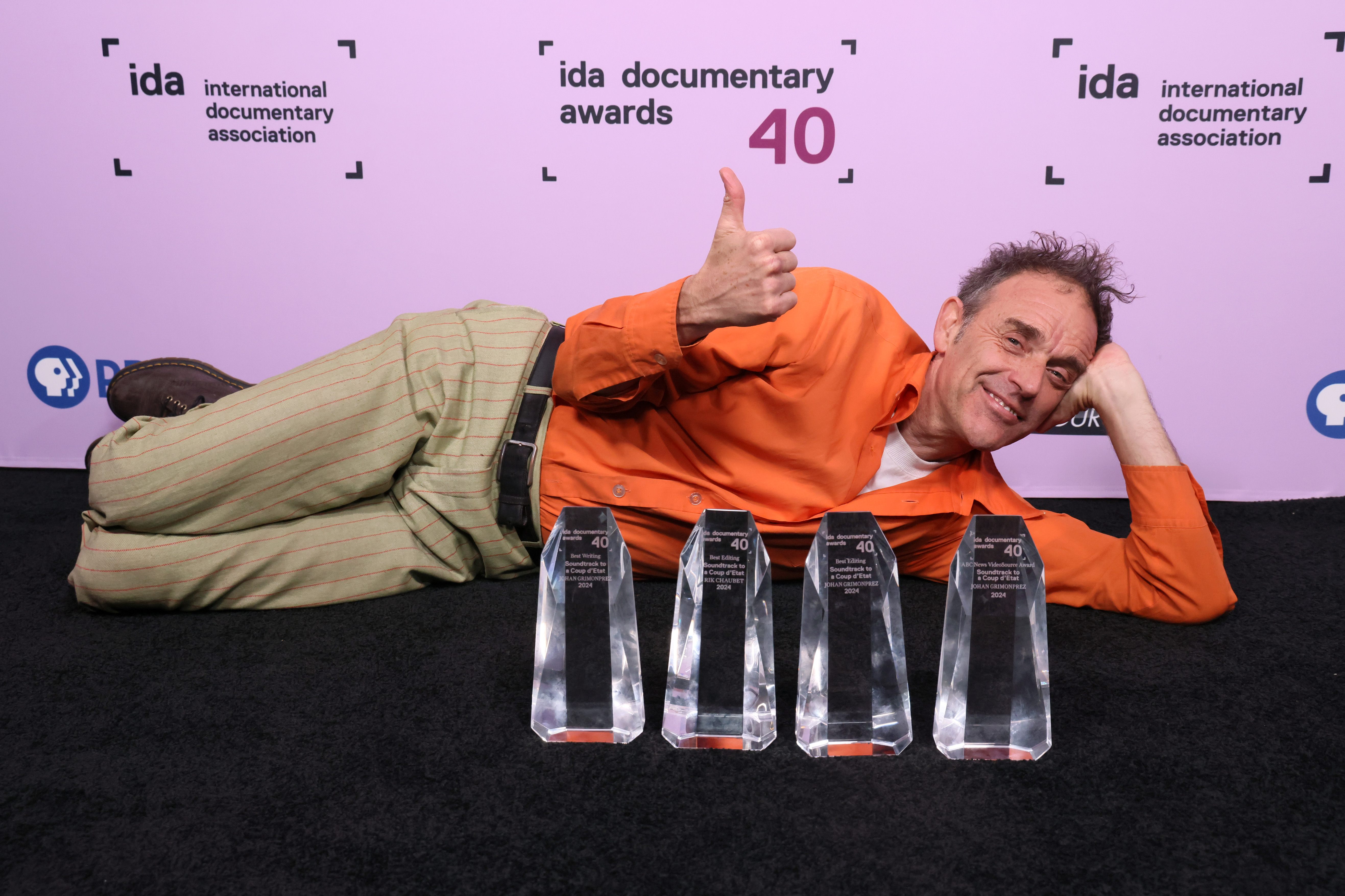 A man in an orange shirt and tan pants lays on the ground with four crystal IDA awards in front of them
