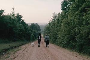 Two women is walking with their white dog on an unpaved road flanked by green green trees.