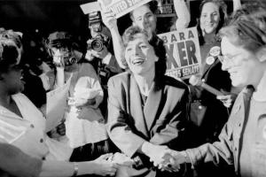 Barbara Boxer is shaking hands with others while supporters hold signs in the background.