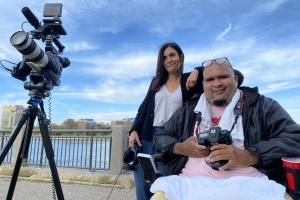 Jay, a Dominican man with a goatee and reading glasses perched on top of his head, sits in a wheelchair holding a digital camera. Alexis, a white woman with dark brown hair stands next to him but a bit behind, holding headphones attached to a large camera on a tripod. They are smiling softly, looking at the camera, and wearing light jackets on a boardwalk in front of a river on a sunny day with clouds in the blue sky.