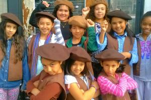A group of young girls of color stand in brown berets with their fists raised. 