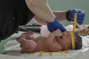A pair of hands wearing medical gloves cares for a crying newborn infant. The nurse is using a tape measure to measure the circumference of the baby's head.