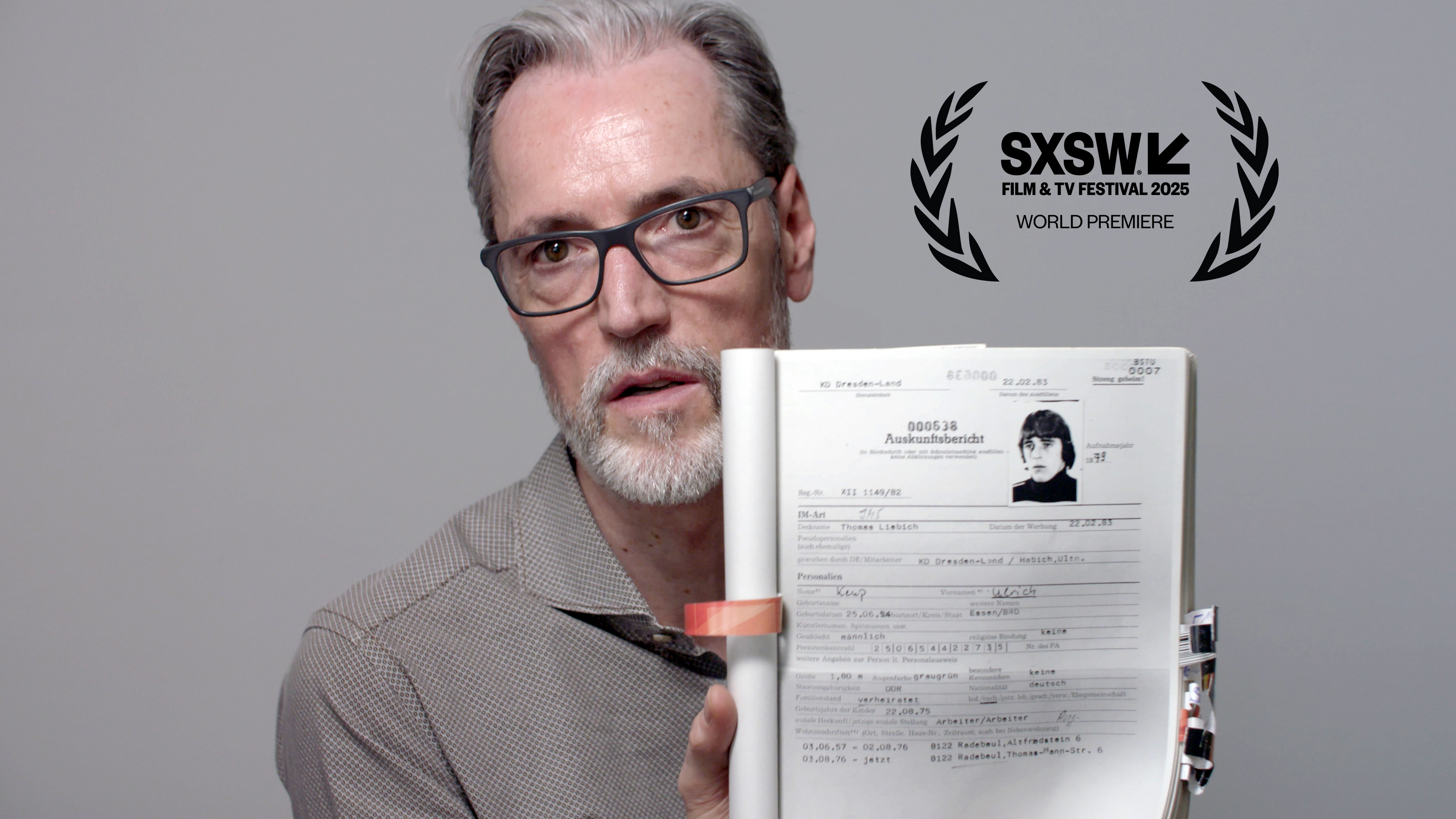 Film still from 'The Spies Among Us via SXSW. A man wearing glasses and a gray button up, holds up a booklet with a black and white picture and data of a person.