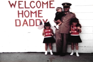 A man in a Navy uniform stands in front of a white garage door, that has "welcome home daddy" written on it in red. The man holds a small child, and two young girls are standing next to him holding American flags.