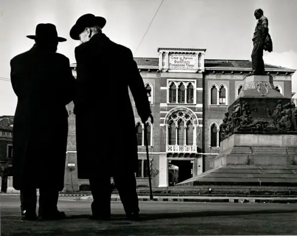 Two men in winter coats and hats in the style of the early 20th century speak to one another closely before a large courtyard with a statue at its center.