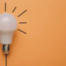 A lightbulb sits on the left side of a soft orange backdrop. Little rays pop out of it, as if they were drawn onto the backdrop. 