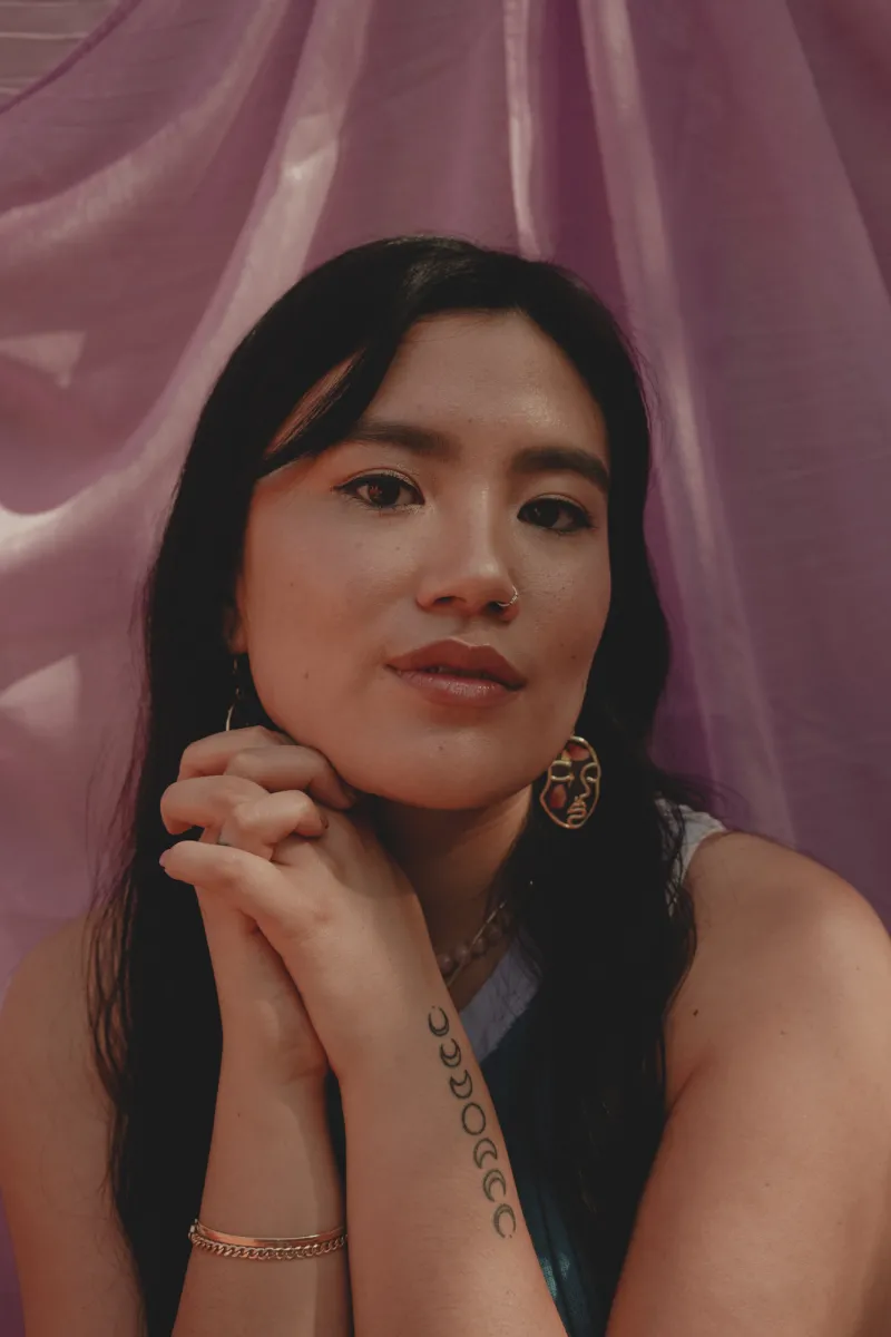 Headshot photograph of Jalena Keane-Lee, whose hands are clasped beneath her chin, and is photographed in front of a dusky pink backdrop.