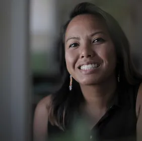 woman with light brown skin and long brown hair smiling at the camera. the background is blurry.